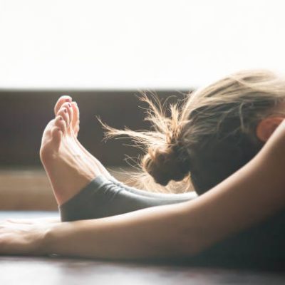 Young woman practicing yoga, sitting in Seated forward bend exercise, paschimottanasana pose, working out, wearing sportswear, grey pants, bra, indoor, home interior background, close up